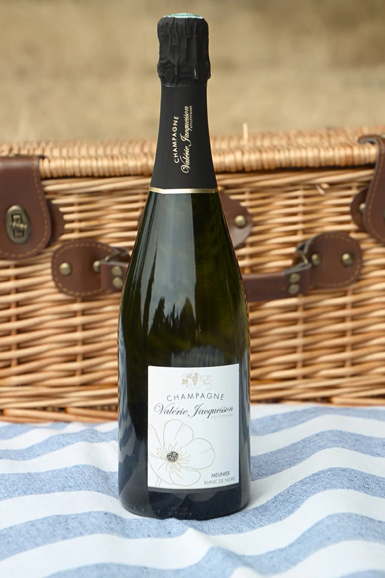 Bouteille de Blanc de Noirs (Meunier) posée devant un panier de pique-nique en osier, sur une nappe rayée bleue et blanche